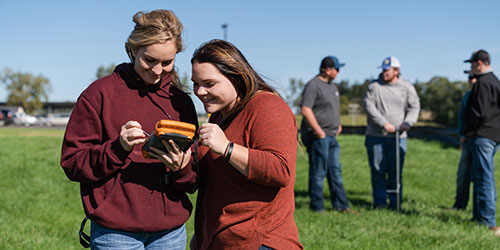 Two female students work with digital reading in foreground while a group of male students work with field samples in background