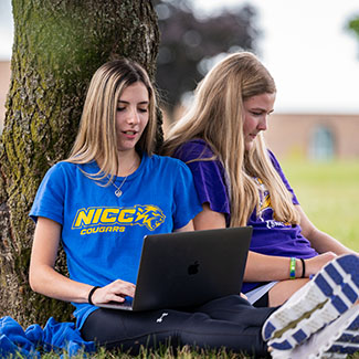 Two NICC students sitting against a tree on the Calmar campus while studying.